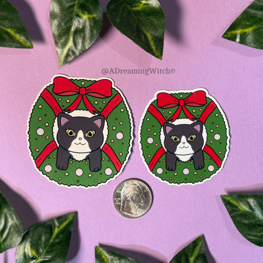 Two stickers of a black and white cat in a green wreath with red bows on a purple background.