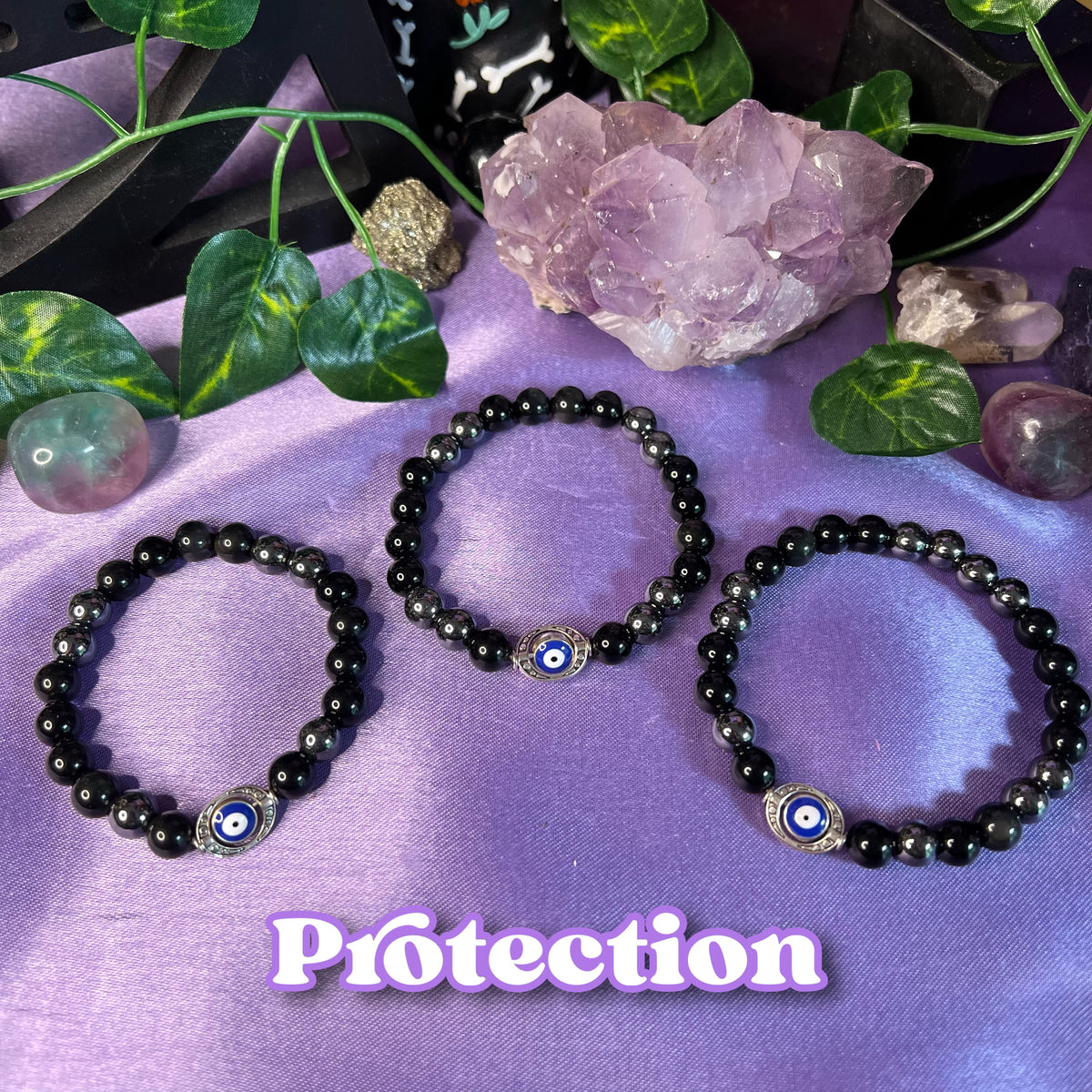 Three black bracelets with different designs, placed on a purple background with a crystal and a leaf, indicating they are protective bracelets with metaphysical properties.