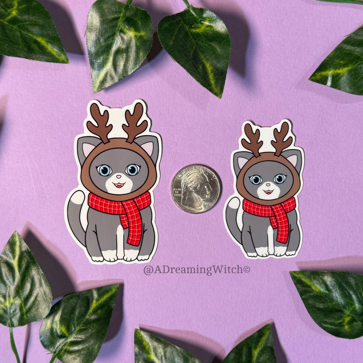 Two stickers of a cat with reindeer antlers and a red scarf on a purple background with leaves.