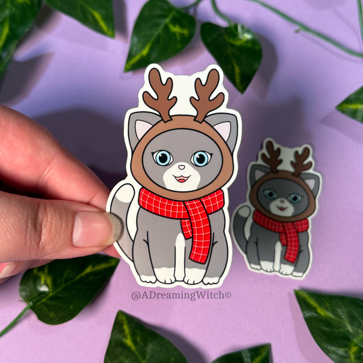Sticker of a cat with reindeer antlers and a red scarf held by a hand on a purple background with leaves.