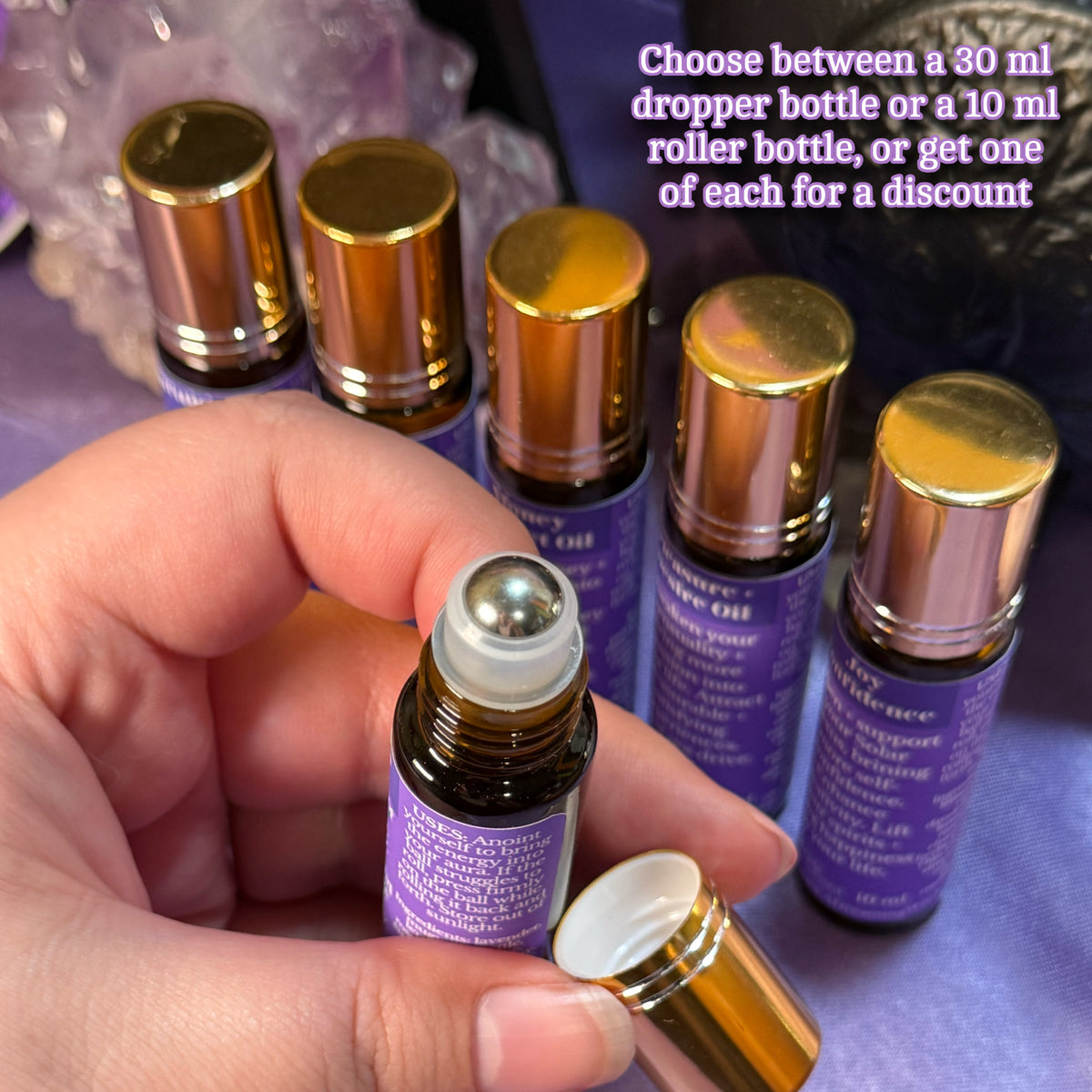 Hand holding a small roller bottle with purple labels and gold caps, surrounded by more bottles on a purple surface.