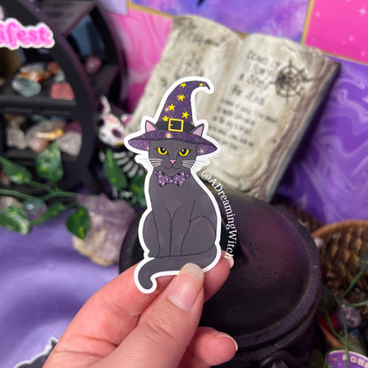 image of hand holding 1 sticker of a black witchy cat, cat wearing a purple witch hat with yellow stars and celestial designs