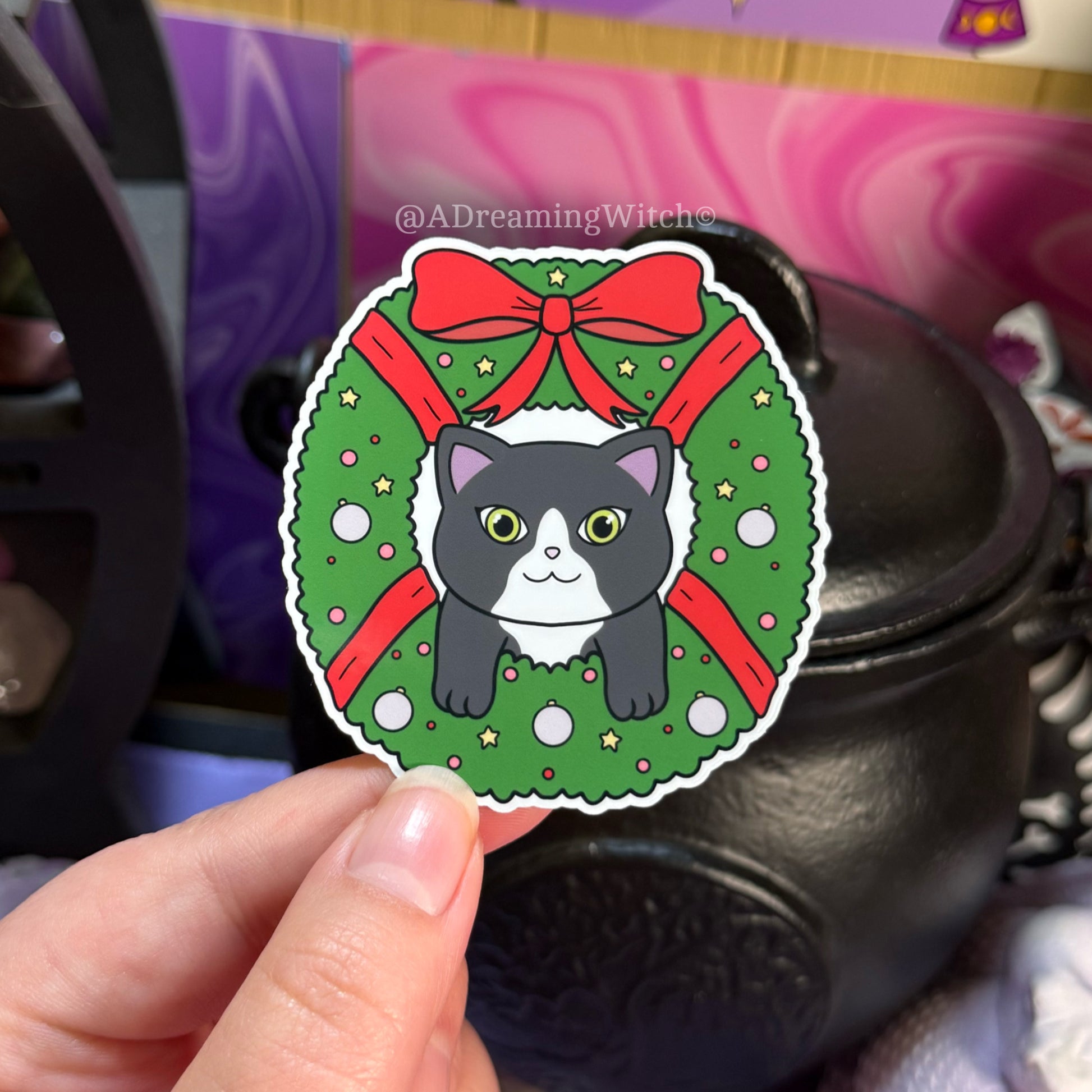 Sticker of a cat in a Christmas wreath held by a hand with a colorful background.