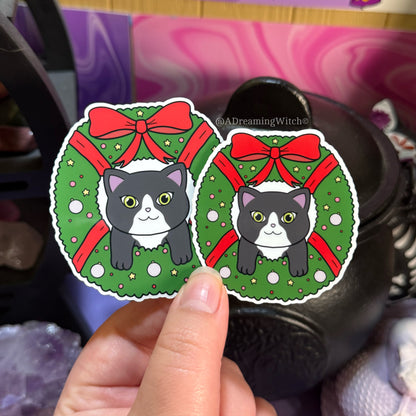 Two stickers of cats in Christmas wreaths held by a hand against a colorful background.