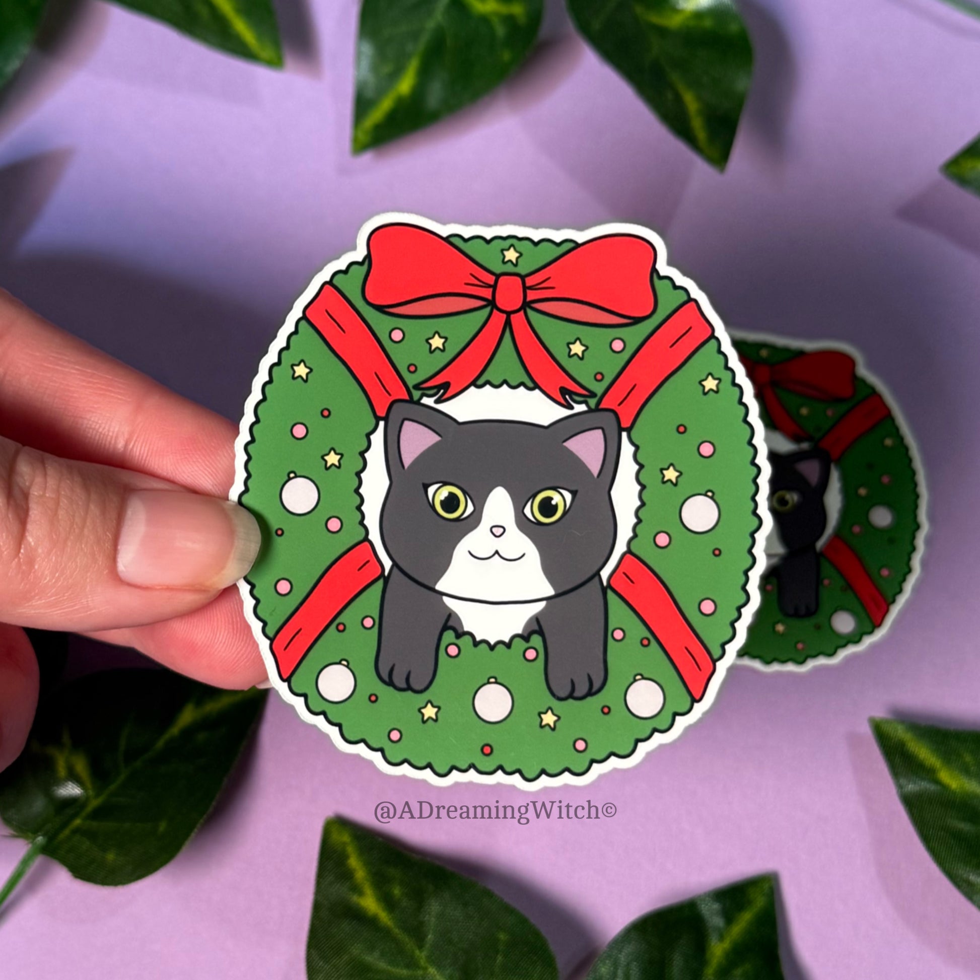 Sticker of a cat in a Christmas wreath held by a hand with green leaves in the background