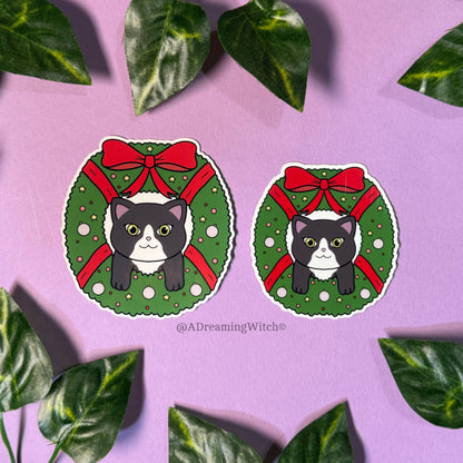 Two stickers of cats in Christmas wreaths on a purple background with leaves.