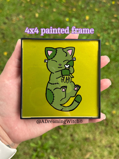 4x4 painted frame with a green Frankenstein cat illustration held by a hand against a grassy background.
