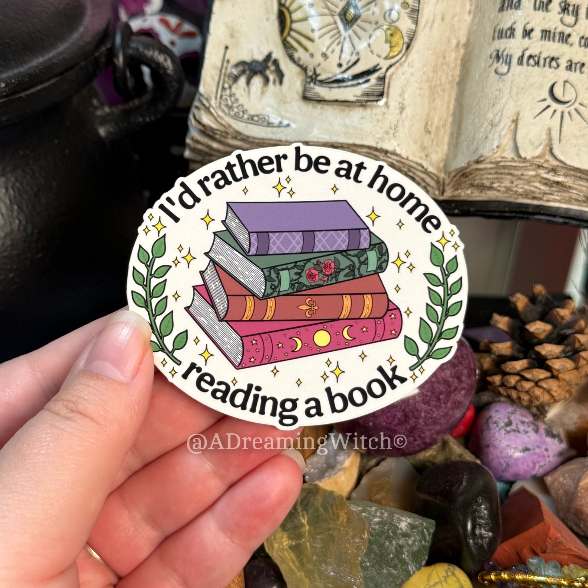 Sticker with books and "i'd rather be at home reading a book" text held in front of a mystical background 