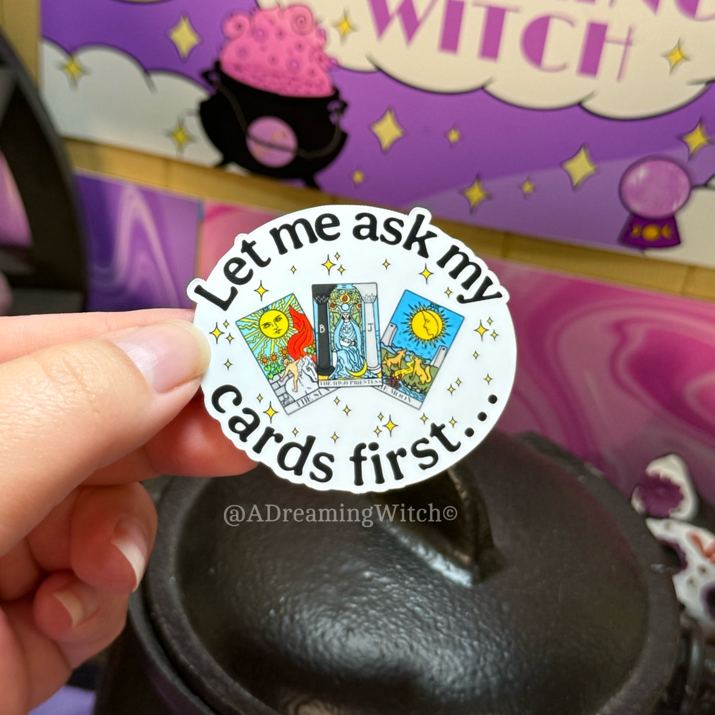 Sticker with tarot card design held by a hand, with a blurred background featuring witch-themed items.