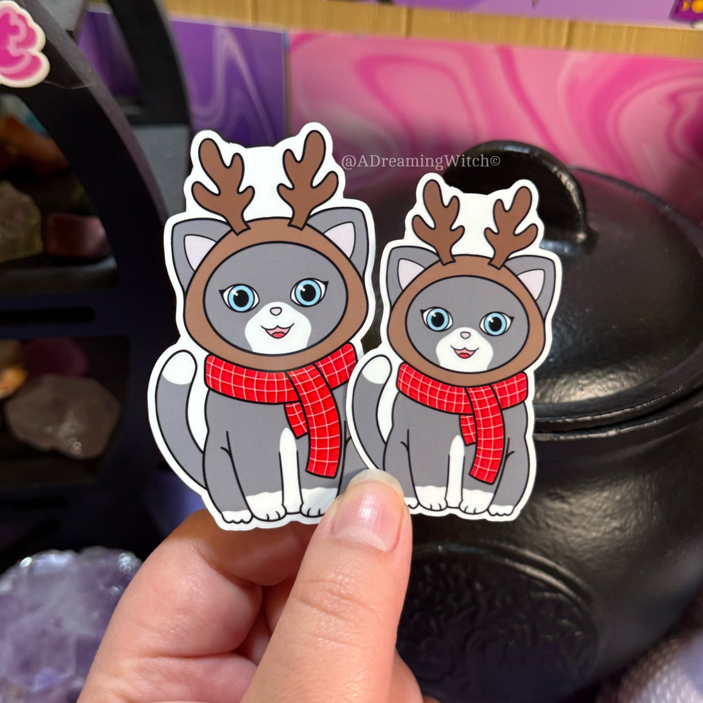 Two stickers of cats wearing reindeer hats and scarves held by a hand.