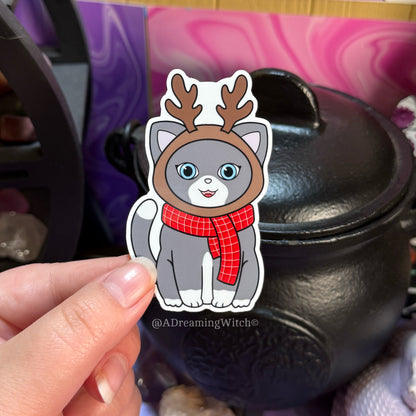 Cartoon cat sticker with reindeer antlers and a red scarf held in front of a black pot.