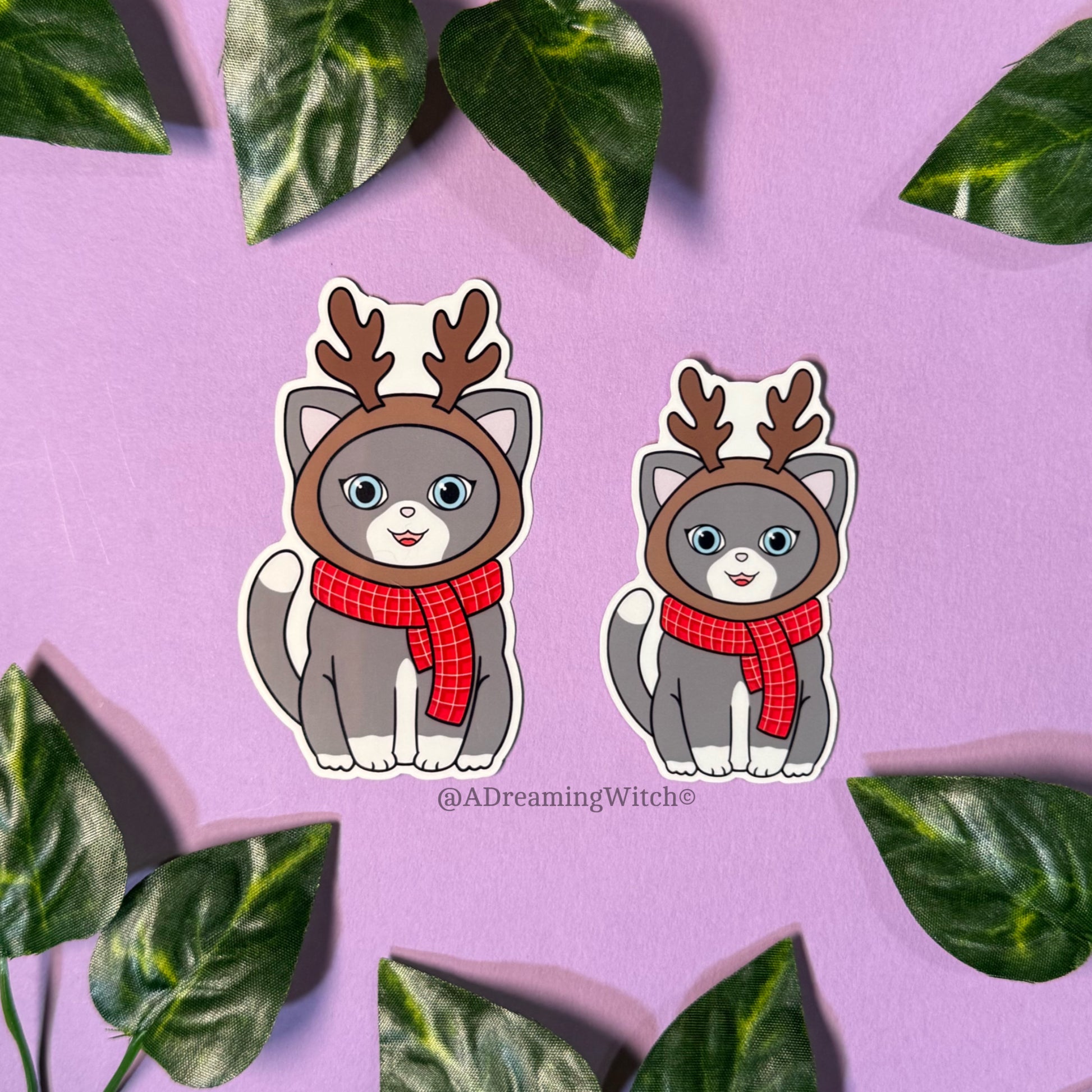 Two cartoon cats wearing reindeer hats and scarves on a purple background with green leaves.