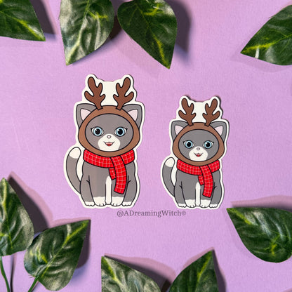 Two cartoon cats wearing reindeer hats and scarves on a purple background with green leaves.