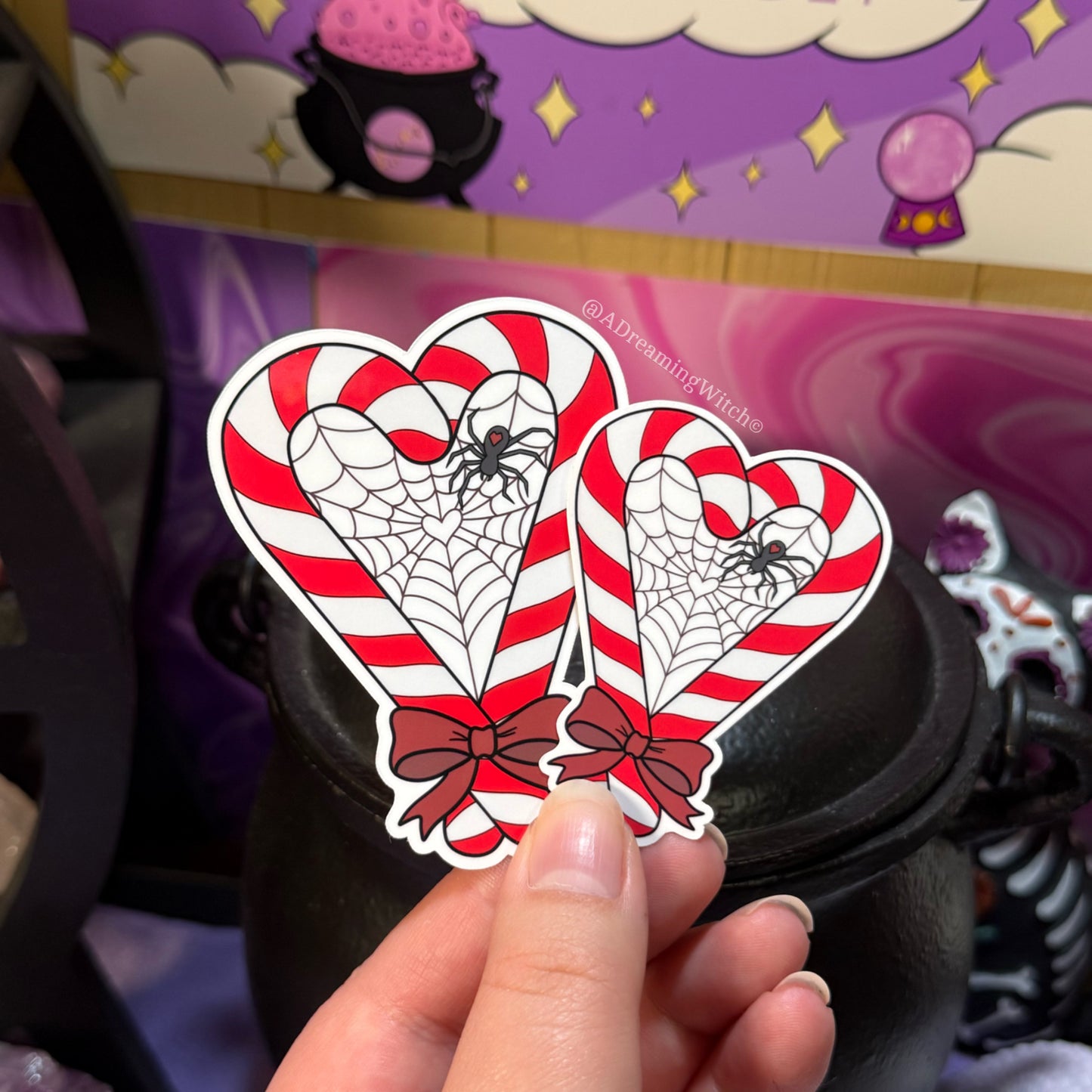 Heart-shaped candy cane stickers with spider webs held by a hand against a purple background with witch-themed decor.