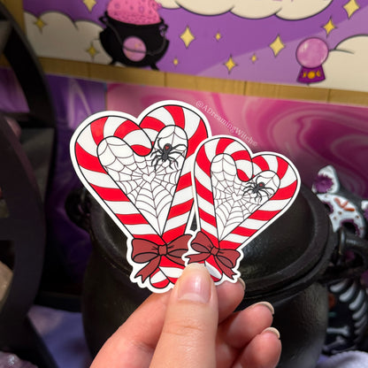 Heart-shaped candy cane stickers with spider webs held by a hand against a purple background with witch-themed decor.