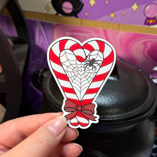 Heart-shaped sticker with candy cane design, spider web, and red bow held by a hand.