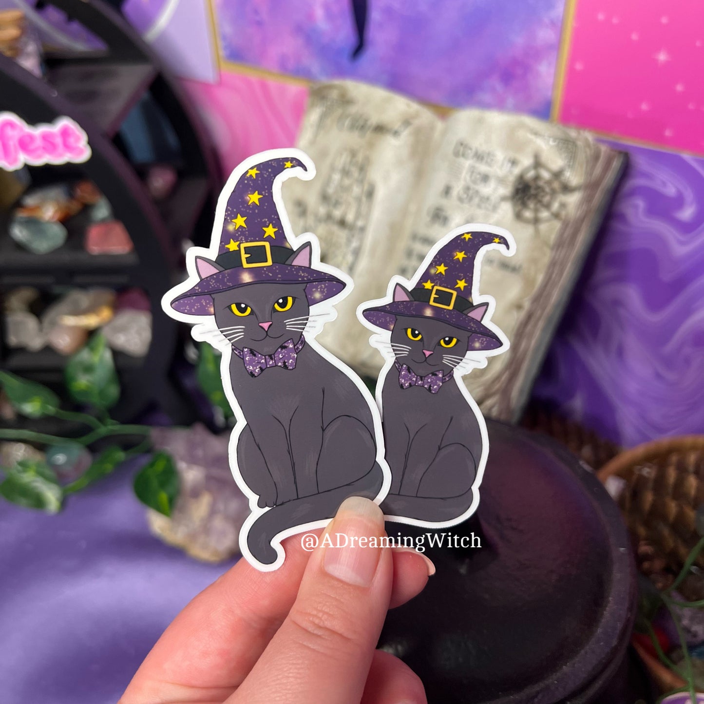 image of hand holding 2 stickers of black witchy cats, wearing a purple witch hat with yellow stars and celestial designs