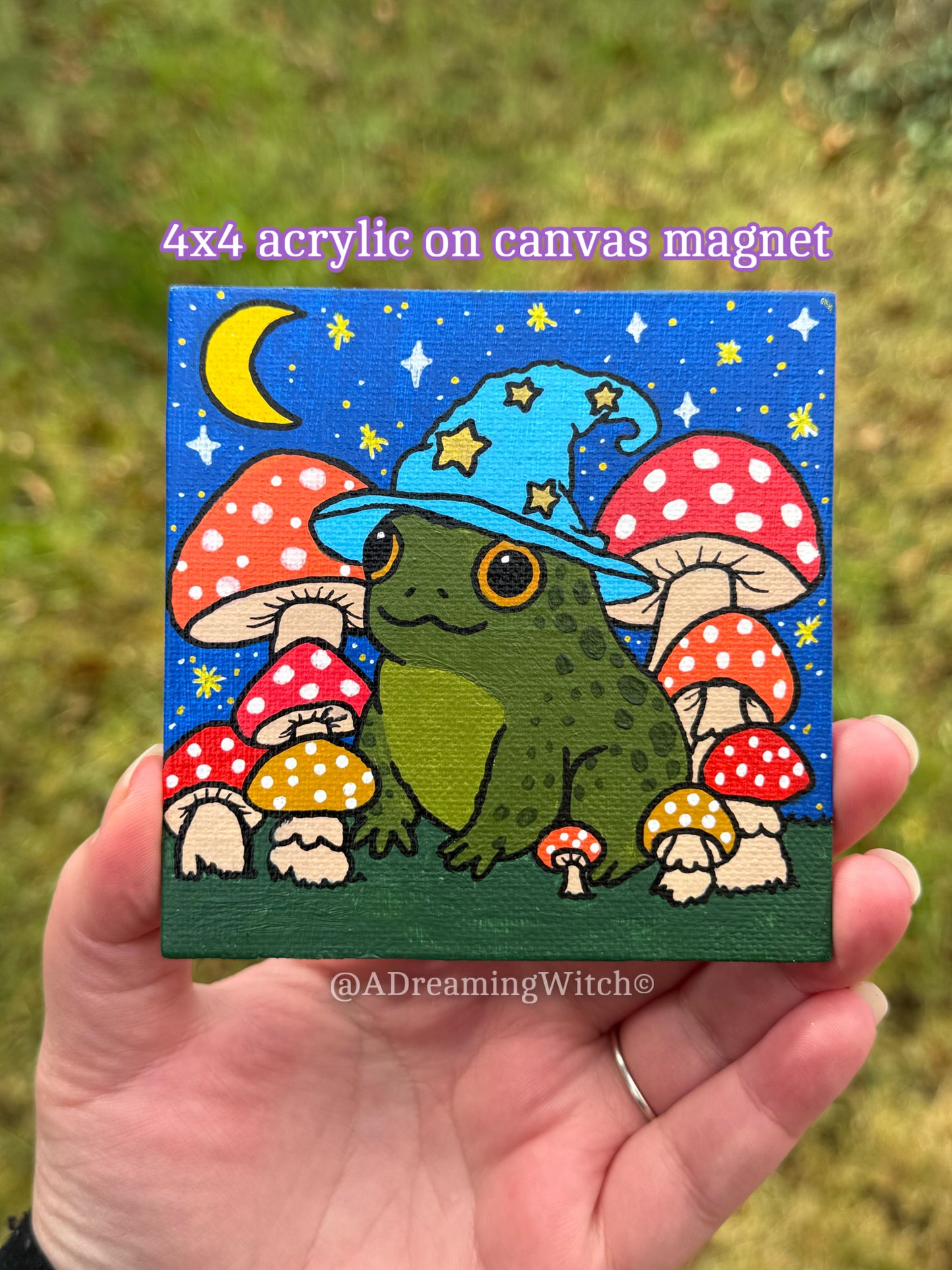 Acrylic on canvas magnet with a frog wearing a witch hat and mushrooms, held in a hand against a grassy background.
